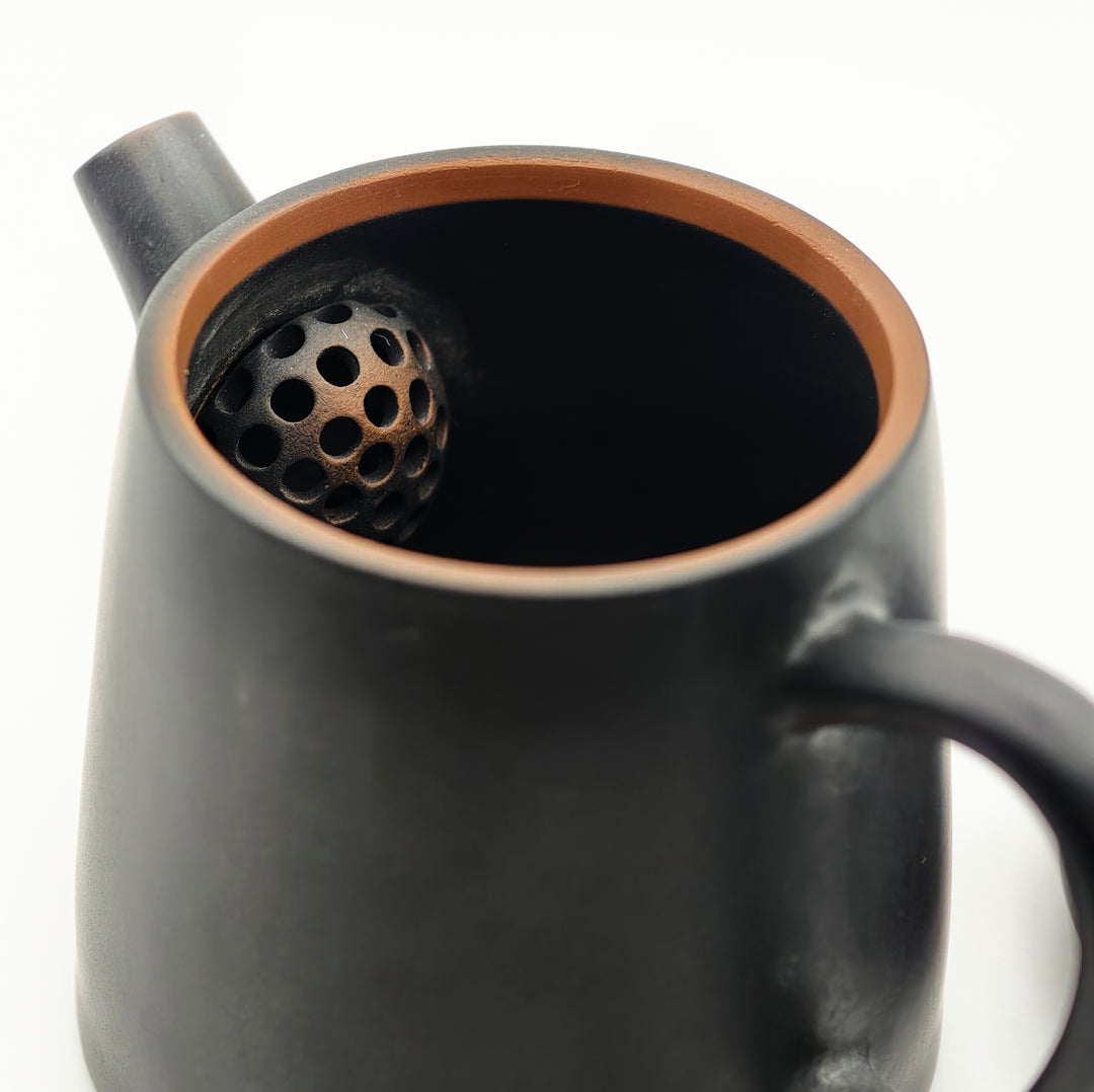 Close-up of the interior of a matte black ceramic teapot with a copper-toned rim, showing a perforated infuser ball inside.