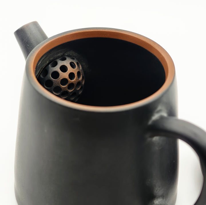 Close-up of the interior of a matte black ceramic teapot with a copper-toned rim, showing a perforated infuser ball inside.